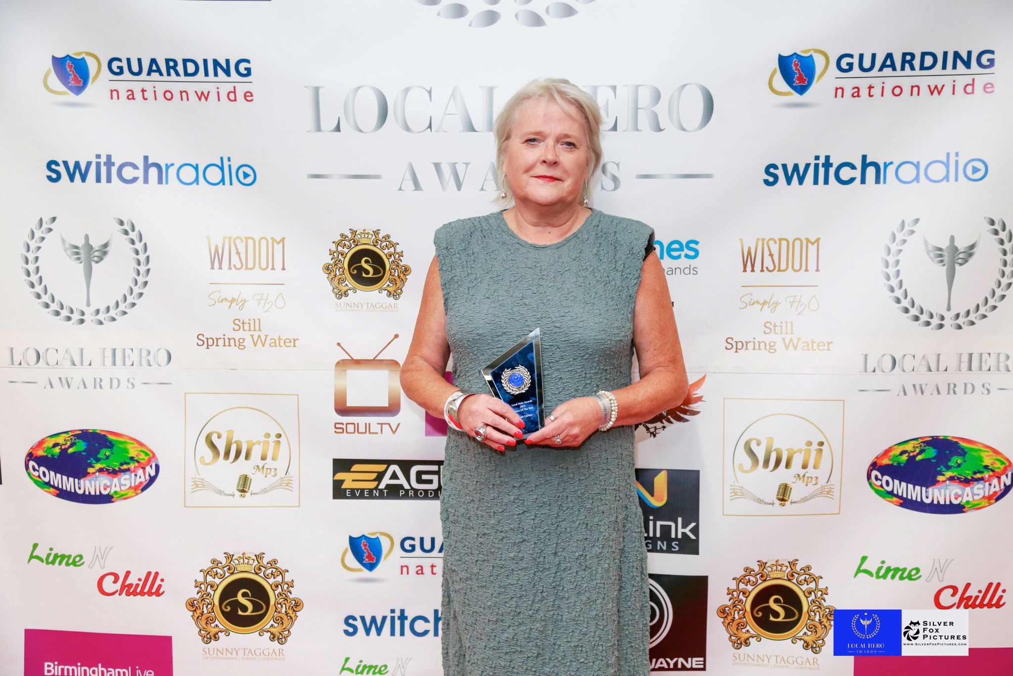 Domestic abuse support worker Ruth Carton holds her award in front of a media board while wearing a cocktail dress.